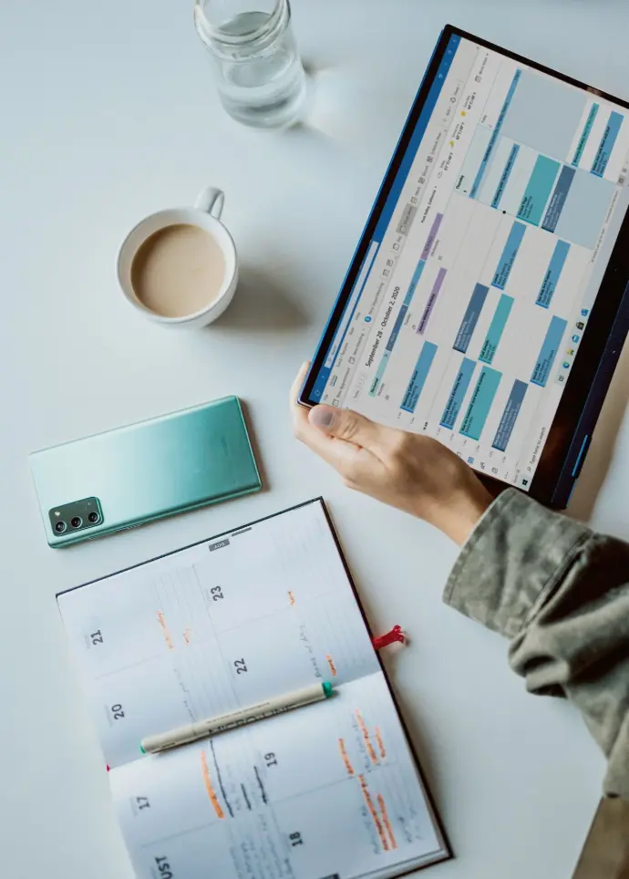 Personne tenant un iPad près d'une tasse en céramique blanche, d'un téléphone portable, d'un agenda papier et d'un ordinateur portable